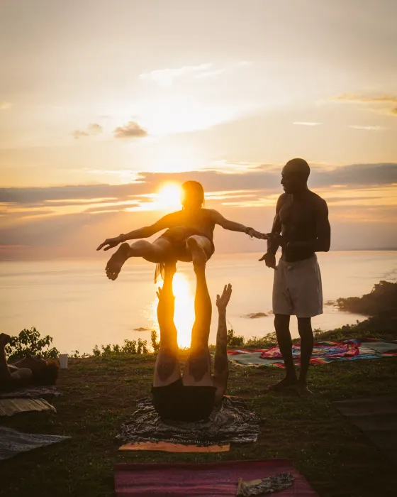 A flyer balancing with arms wide on a partner's feet at Amora, the sun setting into the Pacific behind them.