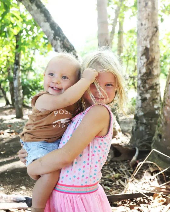 A small girl carrying a laughing baby on her hip in the shade of the trees on the land at Amora.