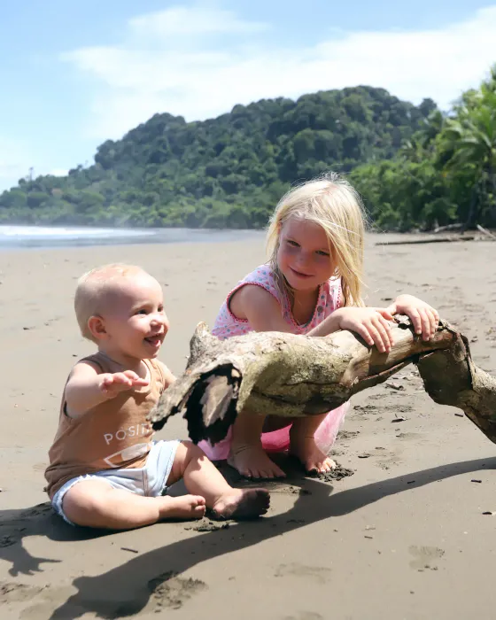 A baby and a young girl playing with a piece of driftwood on the dark sand at the beach below Amora, the forested headland of the Costa Ballena behind.