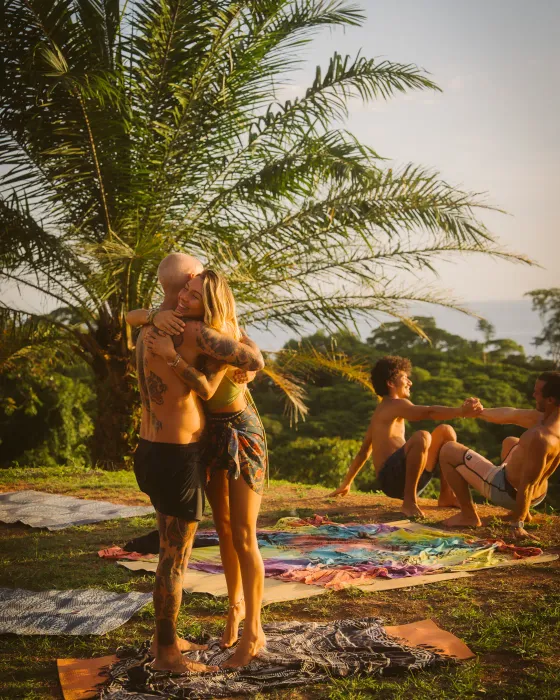 Two friends embracing under the palm after practice at Amora, partners exercising on the grass behind.