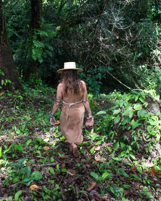 A woman in a straw hat walking a forest path at Amora with a young plant in her hand — reforestation on the land of a regenerative eco village in Costa Rica.