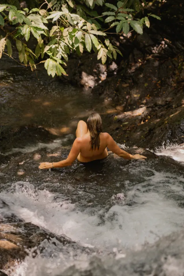 A woman sits in a shallow river pool below whitewater, seen from behind with her shoulders above the surface, in the river that runs through the land at Amora, Dominicalito.