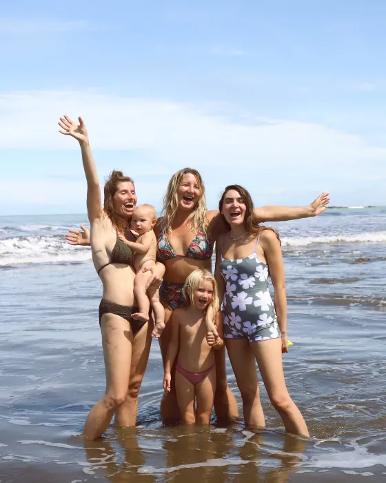 Three women and two small children standing in the surf at the beach below Amora, laughing with their arms thrown wide.