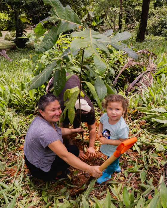 A woman and a small child planting a sapling together, the child holding a trowel, on the land at Amora, a regenerative community near Uvita, Costa Rica.