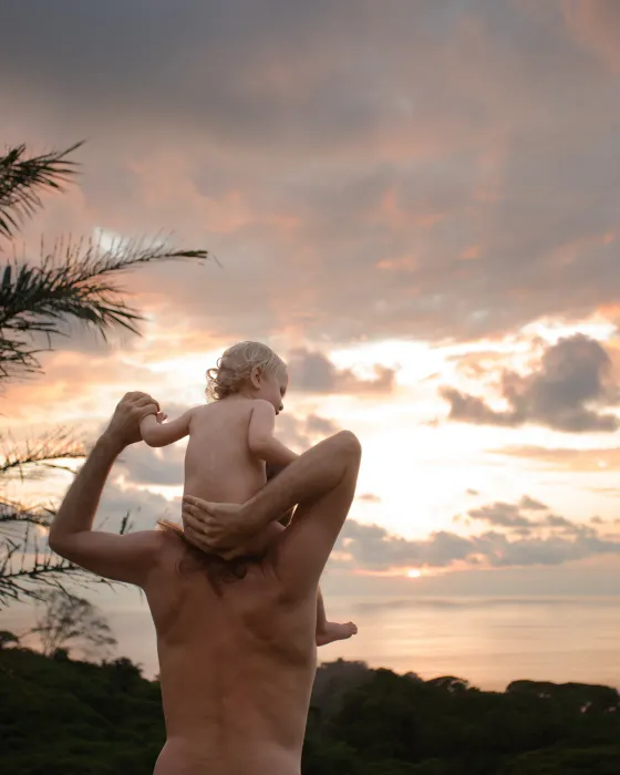 A man with a baby on his shoulders at Amora, both facing a pink and gold sunset over the Pacific.