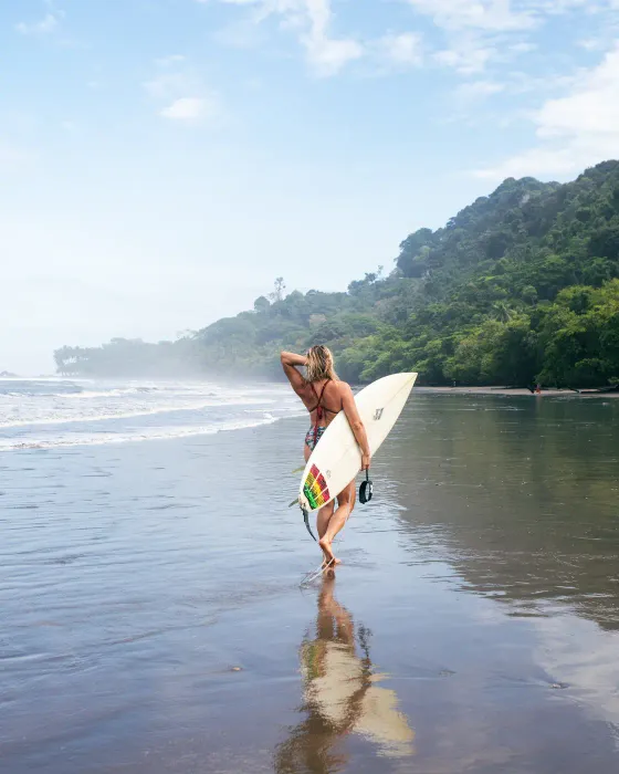 A woman carries her surfboard into the shallows at the beach below Amora, early morning — the eco village sits above the coast near Dominical, Costa Rica.