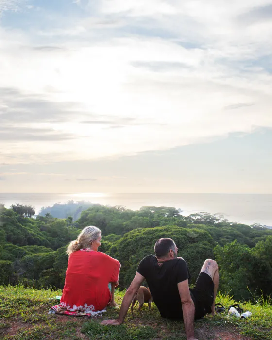 Two people sitting side by side on the ridge grass at Amora, looking out over the forest to the sea — the ocean view from the Costa Ballena ridge, close to Dominical.