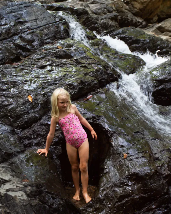 A girl in a swimsuit stepping carefully across the wet rock at the foot of a waterfall on the land at Amora.